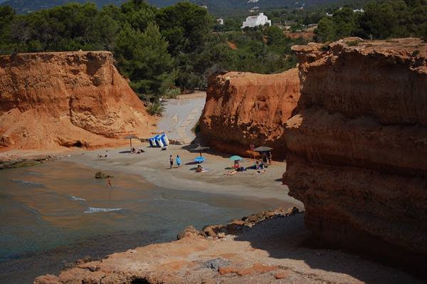 Beachgoers with umbrellas beneath Sa Caleta's red cliffs