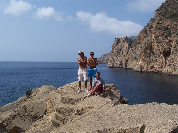Three friends in swimwear pose on rocks beside steep cliffs and blue sea