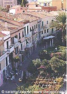 stone-paved street between colorful houses with tiled roofs, palms and pedestrians