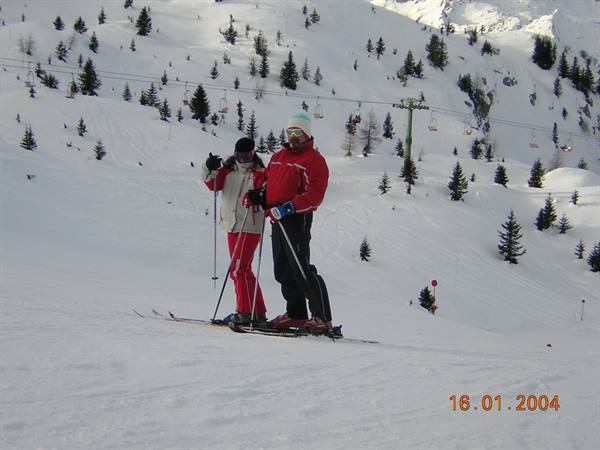 Two skiers pose on snow, chairlift and snowy firs behind them