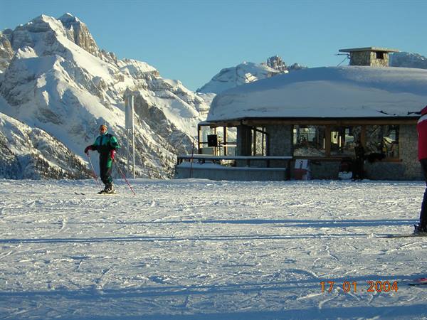 Skier with poles beside a snow-covered mountain hut beneath jagged peaks