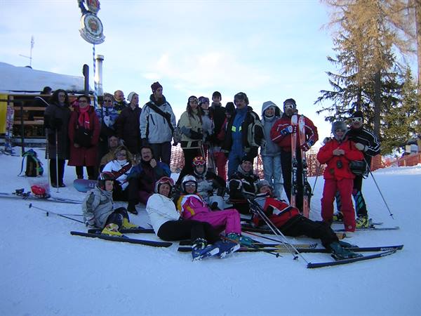 Group of skiers in colorful suits posing on snow in front of a wooden hut