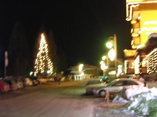 Illuminated Christmas tree on a snowy mountain street at night with parked cars