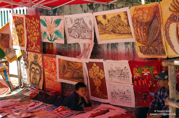 Boy lying under paintings and posters hanging on a shaded market wall