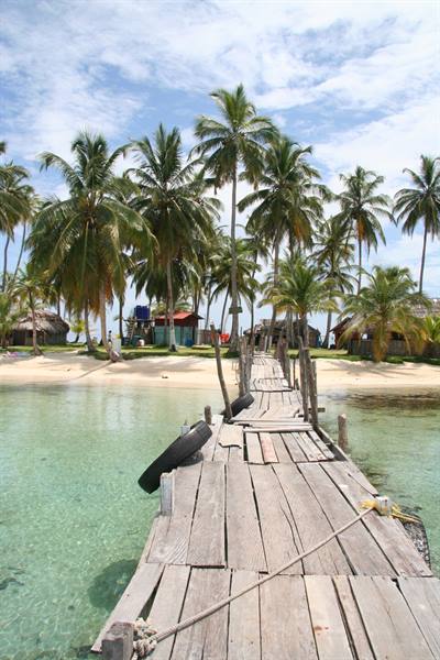 Wooden pier over turquoise water leading to palm-lined beach with huts