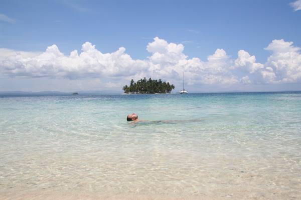 Swimmer in clear turquoise water, palm island and sailboat in the background