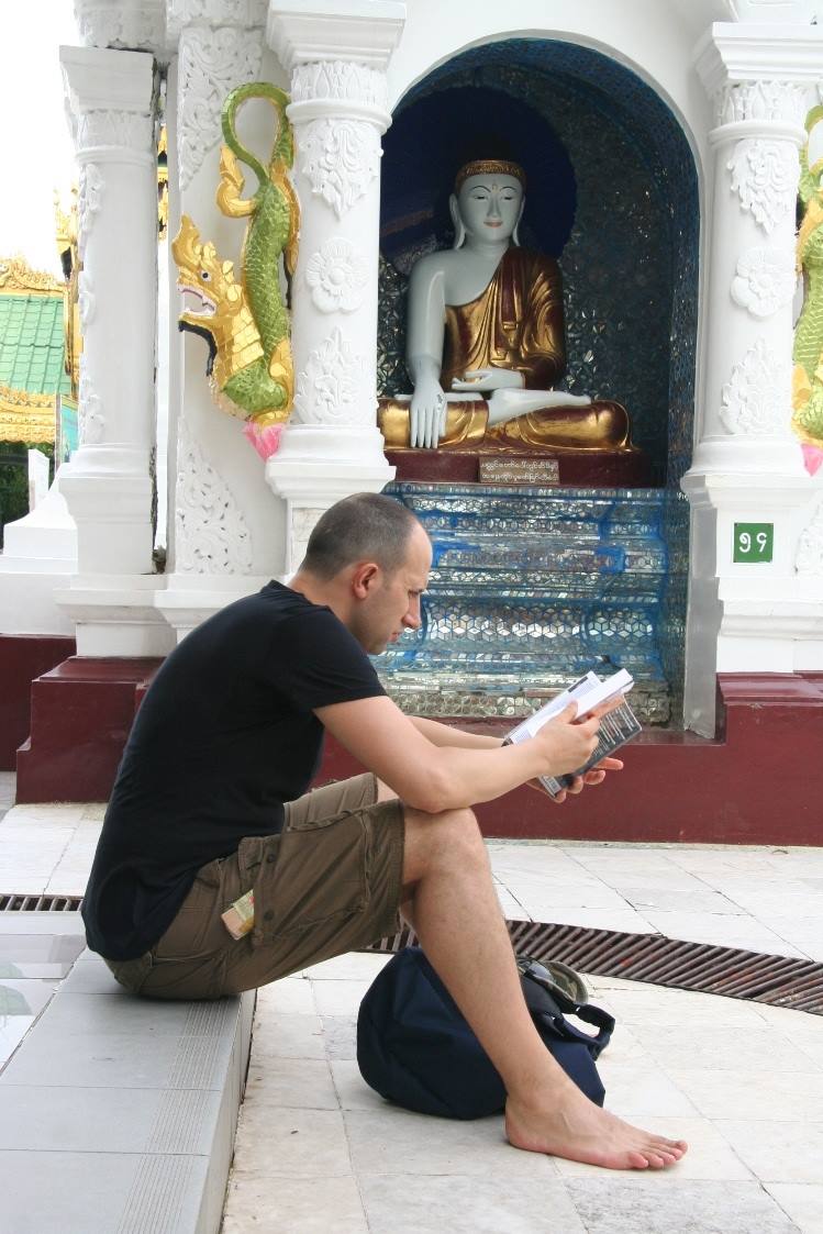 Uomo seduto scalzo legge un libro accanto a una statua di Buddha dorata nella pagoda