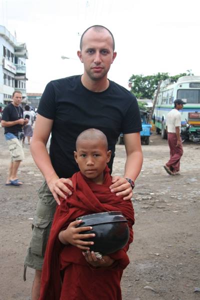 Tourist stands behind young monk in red robe holding black bowl, busy street