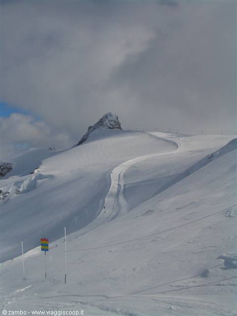 Snowy ski slope winding between rocky peaks under grey clouds
