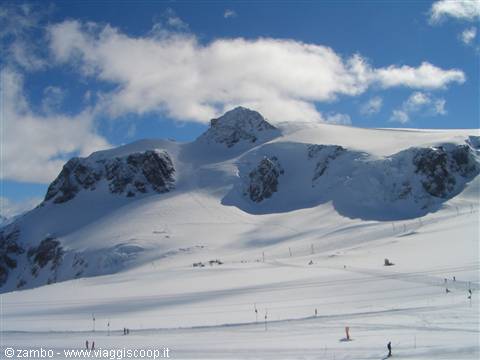 Skiers on snowy slope beneath Presena glacier rocks, cloudy blue sky