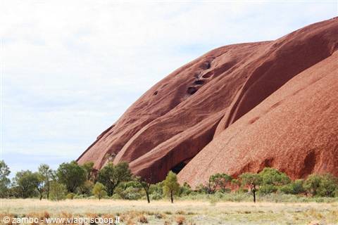 Red sandstone monolith of Uluru rising above scrubland under cloudy sky