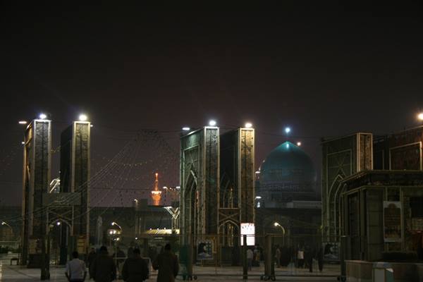 Night at Mashad shrine, green dome glowing among decorated gateways and visitors