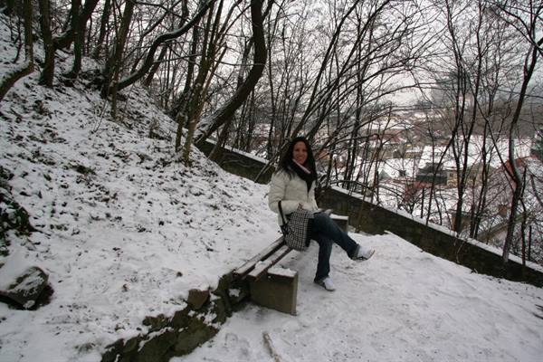 Smiling woman sitting on snowy wooden bench, bare trees and rooftops around