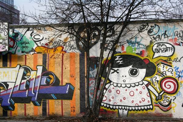 Graffiti wall with yellow-hooded cartoon doll, bare tree in front, cold light
