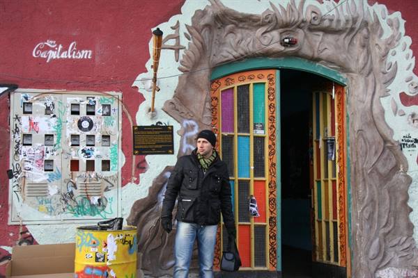 Man at colorful door of Metelkova, dark sculptures and graffiti writings