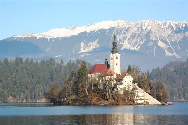 Church with bell tower on Bled Island, snowy mountains and fir trees