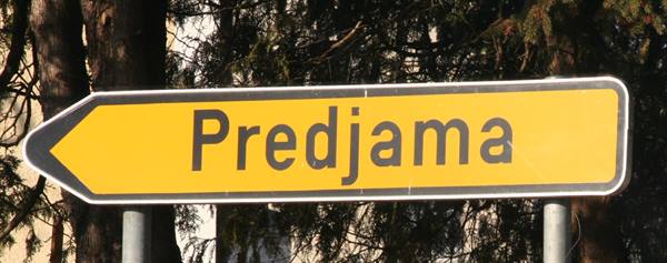 Yellow road sign with arrow pointing to Predjama, among backlit trees
