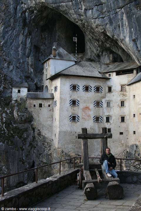 Man sitting by a wooden cart in front of Predjama Castle, built into the rock face