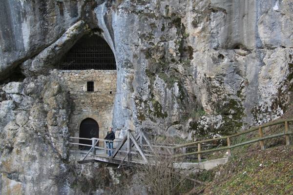 Stone building built into cliff with barred cave entrance, tourist on wooden bridge, Predjama