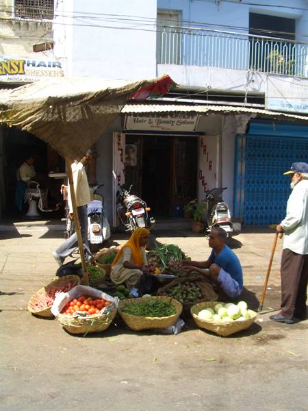 Vegetable vendors squatting among baskets of tomatoes and cabbages under straw parasol