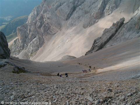 Climbers descend a steep scree slope beneath grey rocky walls