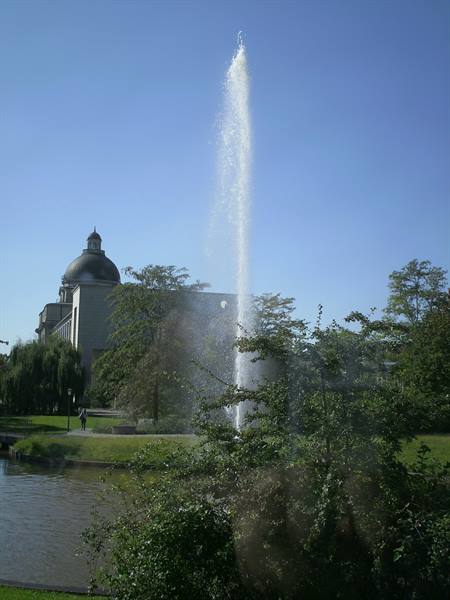 Tall water jet over pond, grey dome behind trees, clear blue sky