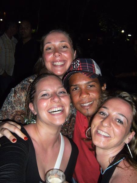Four friends smiling together at night, one wearing red cap, holding a drink