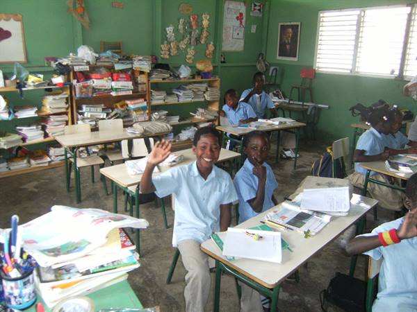 Students in light blue uniforms wave and smile among desks and books, green classroom walls