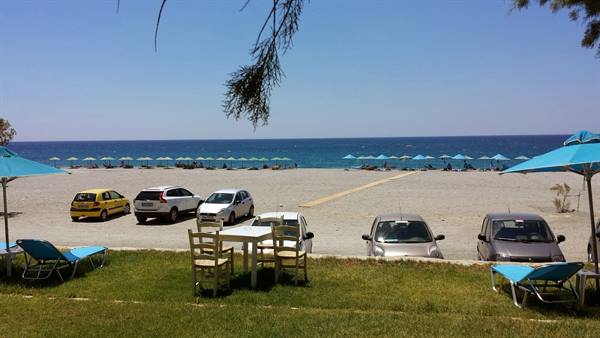 An empty little table on the grass, in front of the beach full of blue umbrellas.