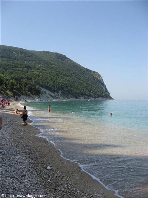 Swimmers on pebbly beach, turquoise sea, green mountain under clear sky, Portonovo