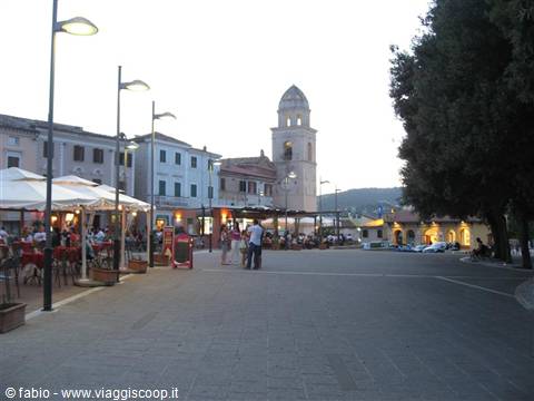 Stone-paved square with cafés under umbrellas and lit bell tower at dusk