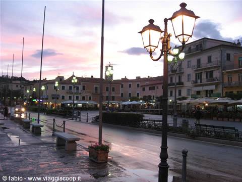 Waterfront square at dusk: lit lamppost, benches, illuminated buildings and pink sky