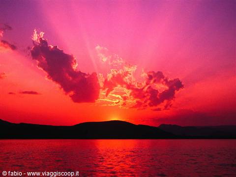 Sun sinking behind a dark hill, vivid pink sky reflecting red over the sea