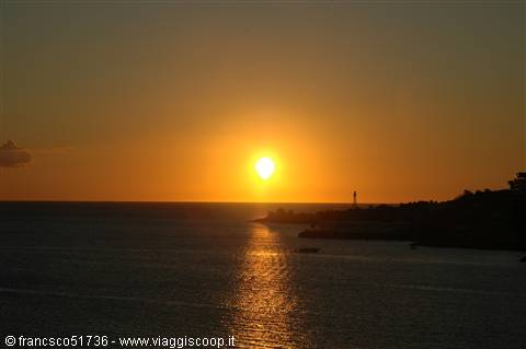 Low sun sinking into the sea behind a lighthouse on the dark coast