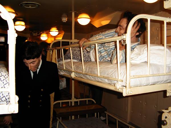 Visitors among metal bunk beds under yellow lamps, sailor mannequin