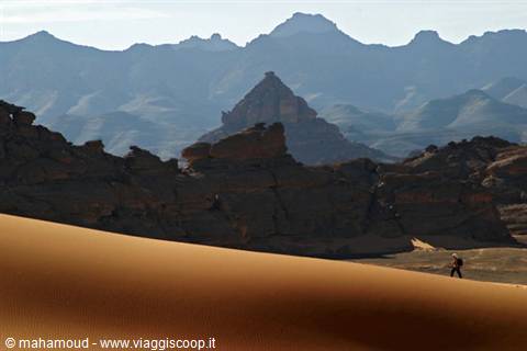 Lone walker on golden dune before dark rocks and blue mountains