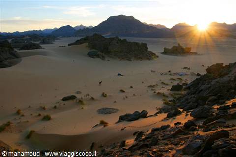 Low sun over black rocks and sandy dunes, a camel in the distance