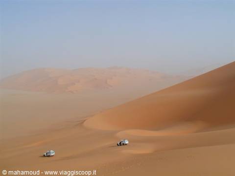 Two white off-road vehicles climb huge orange sand dunes