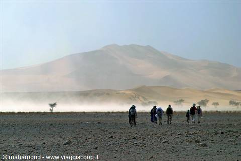 Hikers walk across rocky ground toward dust-covered sand dunes