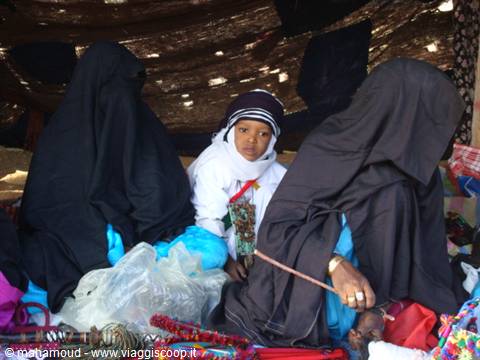 Child between two black-veiled women at shaded market, colorful goods scattered