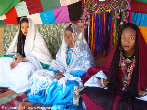 Tuareg women in white and blue veils seated under colorful fabrics