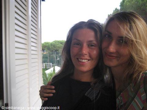 Two smiling young women embracing on a balcony beside white shutters