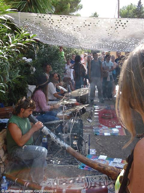 Musician plays the didgeridoo under a shade net, crowd watching the drummers