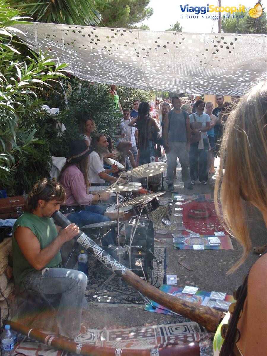 Musician plays the didgeridoo under a shade net, crowd watching the drummers