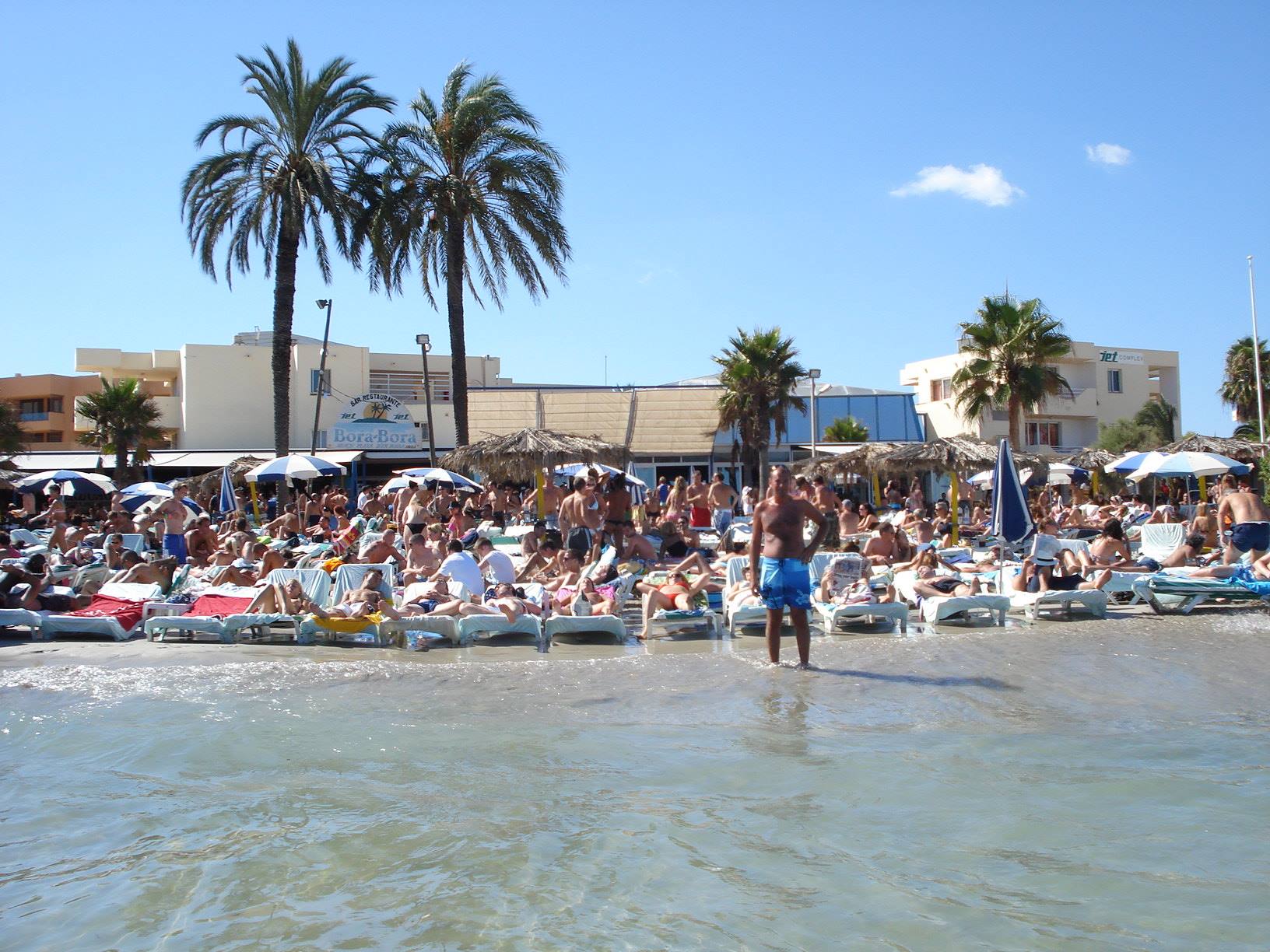 Crowded beach at British Bath: people on white sunbeds, palms and umbrellas, man wading
