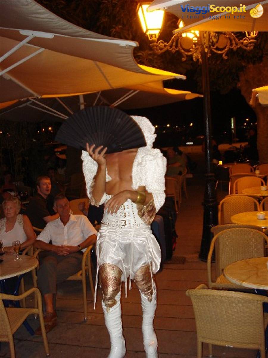 Drag queen in white and gold with black fan among night café tables
