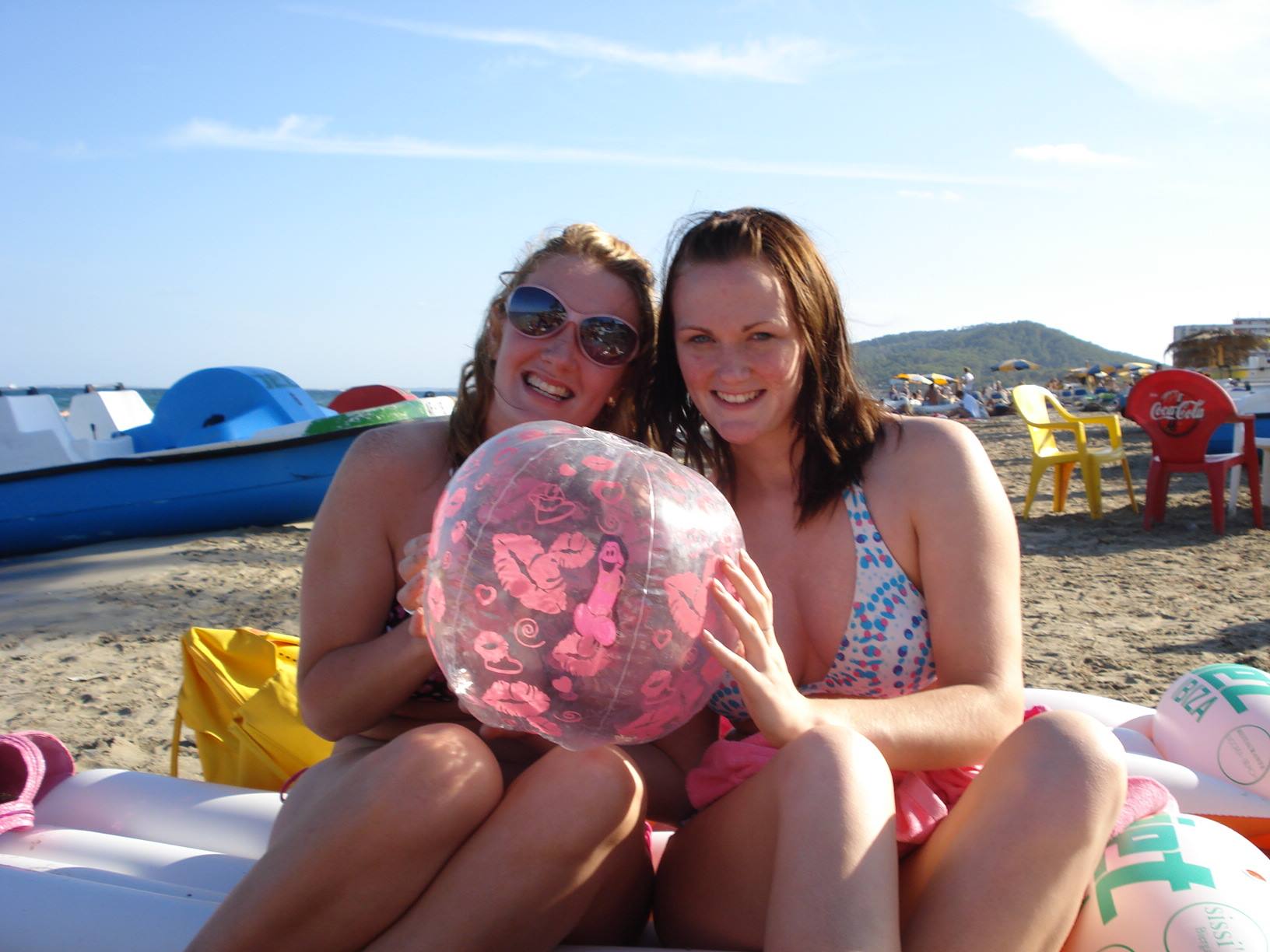 Two girls in swimwear smiling on the beach holding a transparent pink balloon