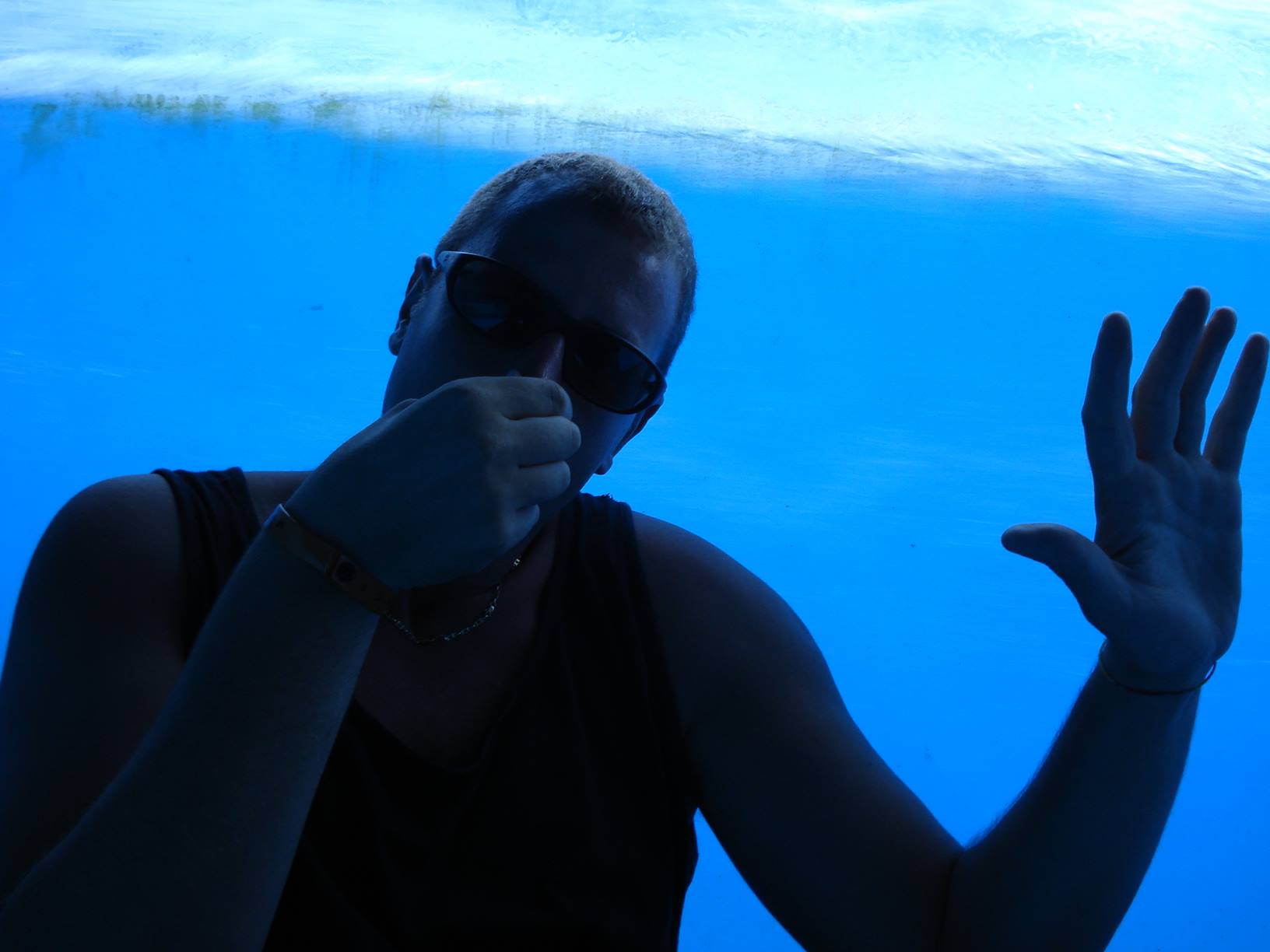 Man in sunglasses and tank top pinching his nose underwater in a pool