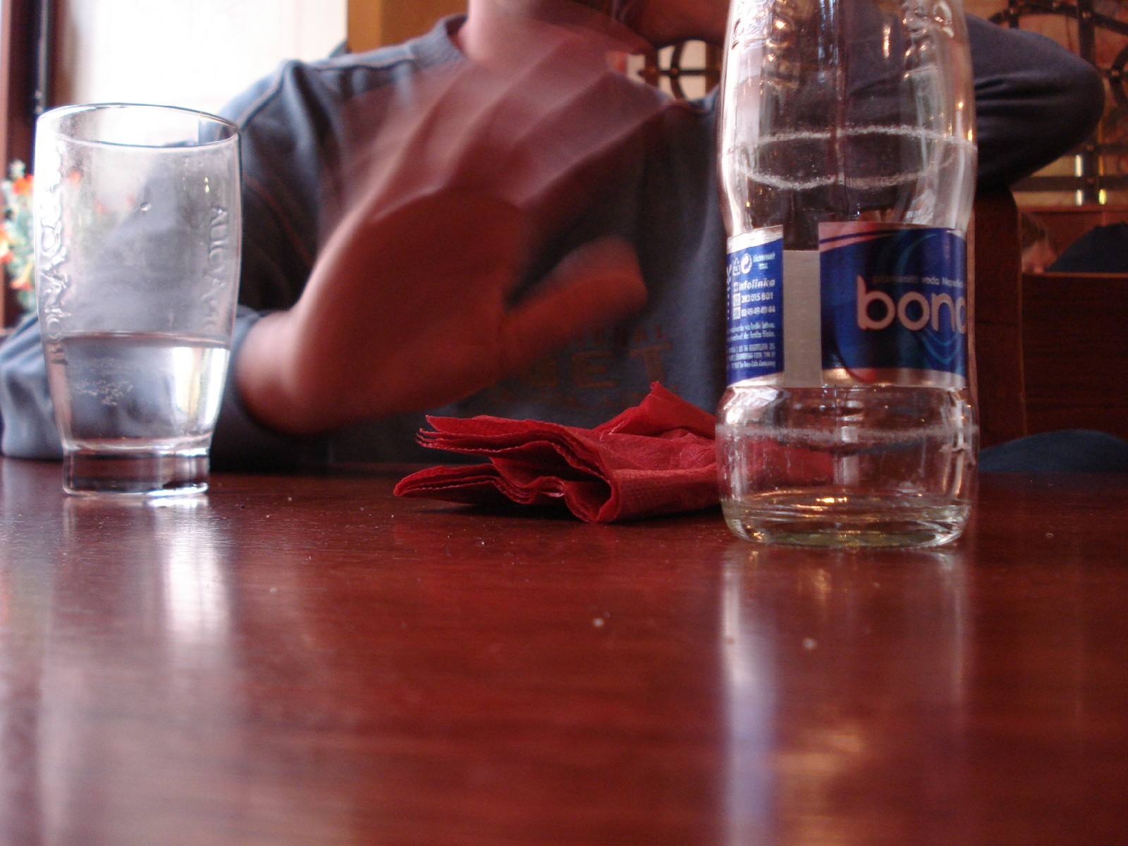 Glass, red napkin and Bon Aqua bottle on wooden table, blurry man behind