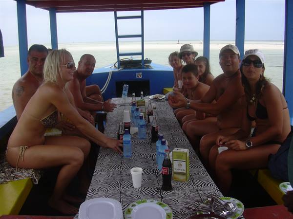 Tourists in swimwear sitting on blue boat before white beach, toasting